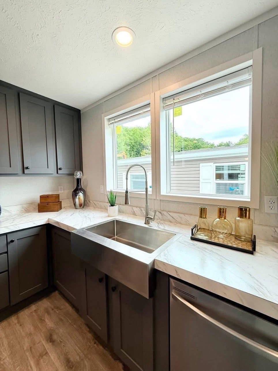Modern kitchen with gray cabinets, marble countertops, and a stainless steel sink. Large windows overlook outdoors, creating a bright, airy feel.