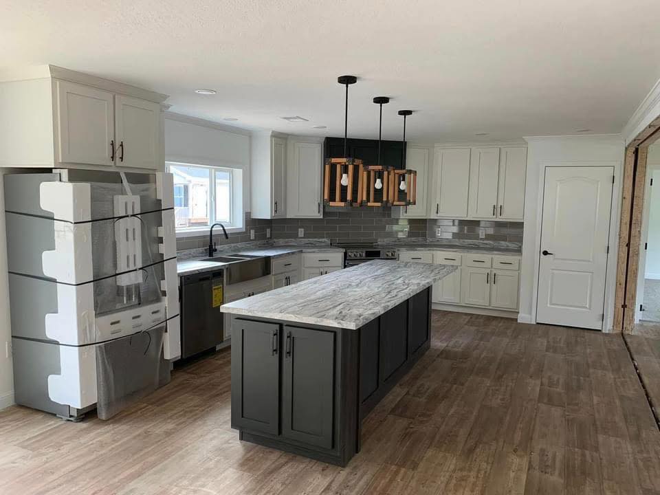 Modern kitchen with gray cabinets, marble countertops, and a central island. Wooden pendant lights hang above; appliances are new and wrapped.
