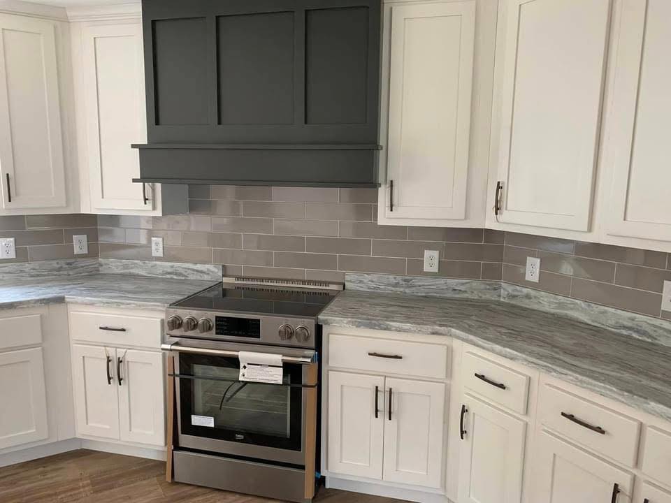 Modern kitchen with white cabinets, gray subway tile backsplash, stainless steel oven, and marbled countertops under a dark gray range hood.