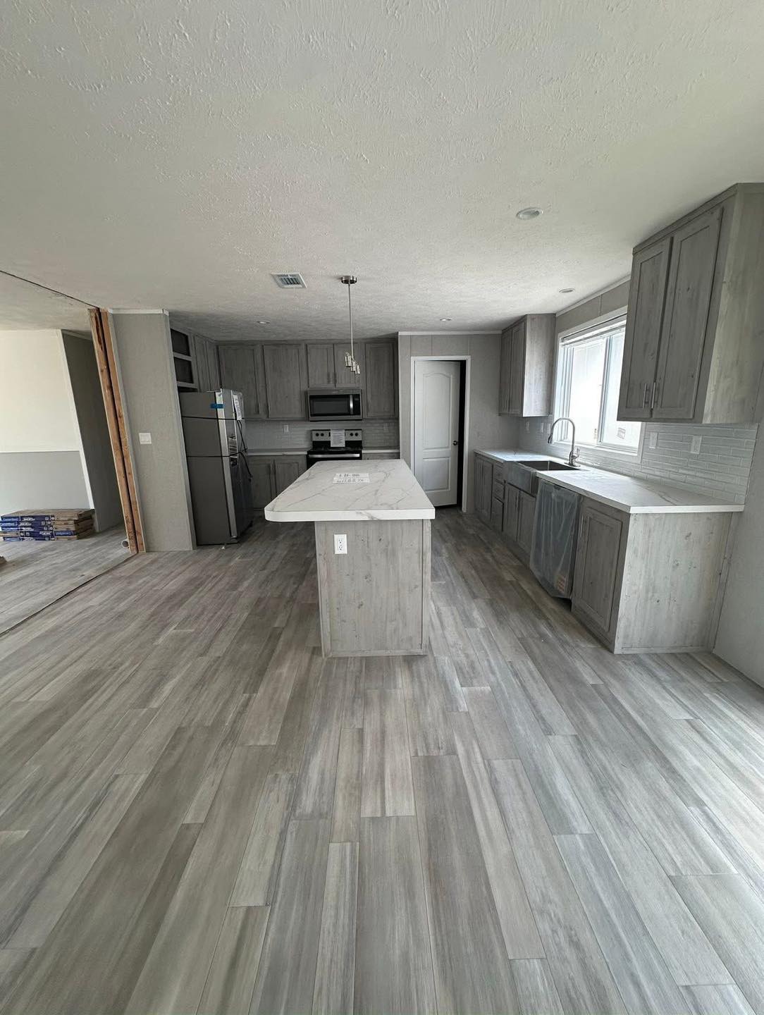 Modern kitchen with light gray wood cabinets, stainless steel appliances, and a marble island. Sunlight streams through a large window, creating a bright and inviting space.