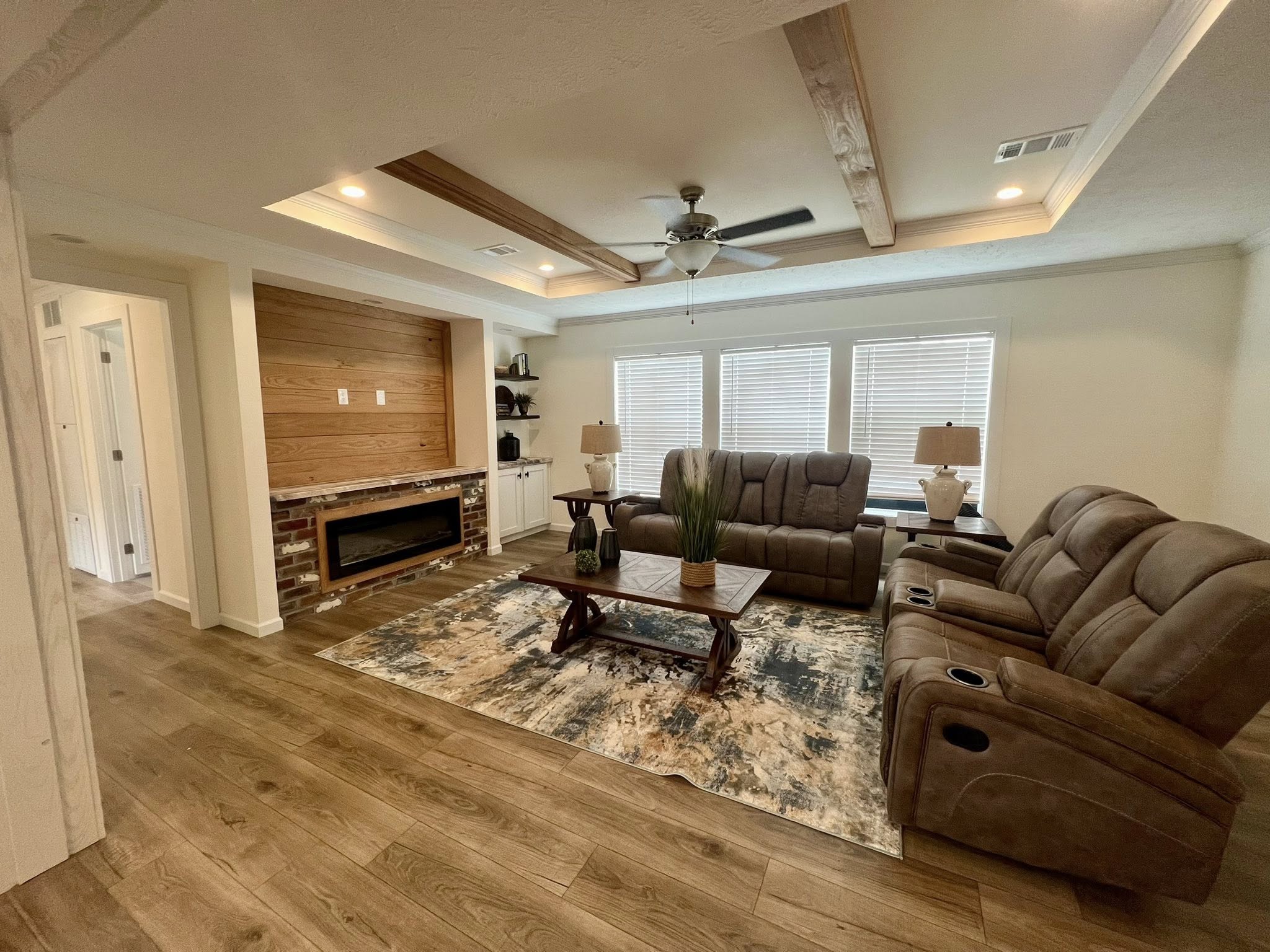 A cozy living room with brown recliners, a wooden coffee table, and a patterned rug. A modern fireplace and large windows add warmth and light.