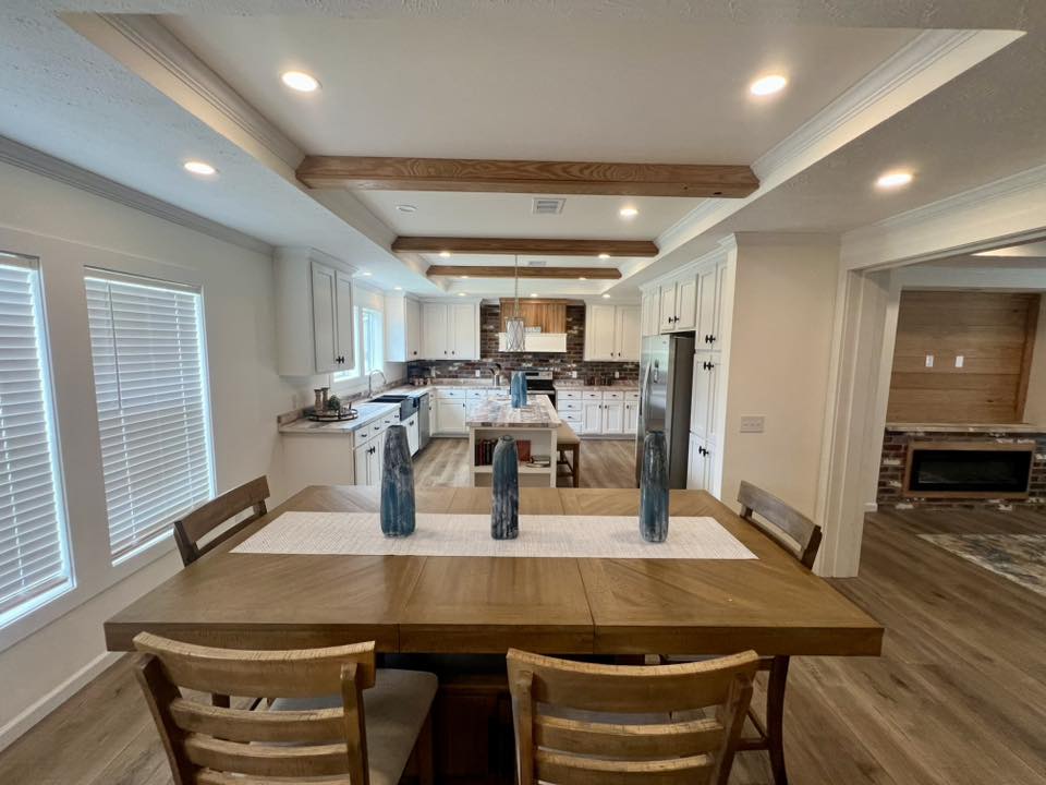 Modern kitchen-dining area with wooden table, white runner, and three blue vases. Bright lighting, white cabinetry, and exposed beams enhance warmth.