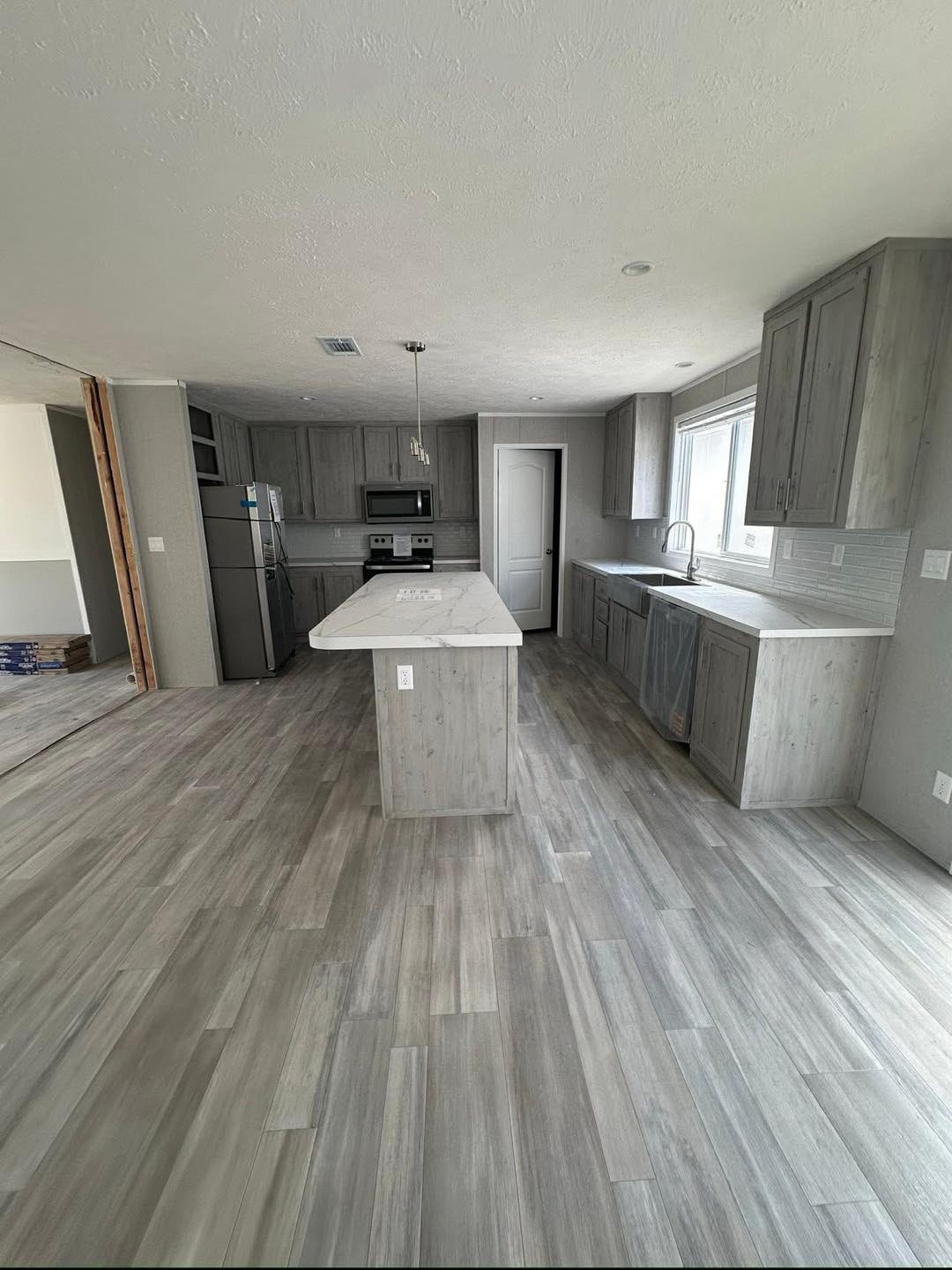 Modern kitchen with sleek gray cabinetry and wood-like flooring. Features a central island, stainless steel appliances, and a large window, giving a bright, open feel.