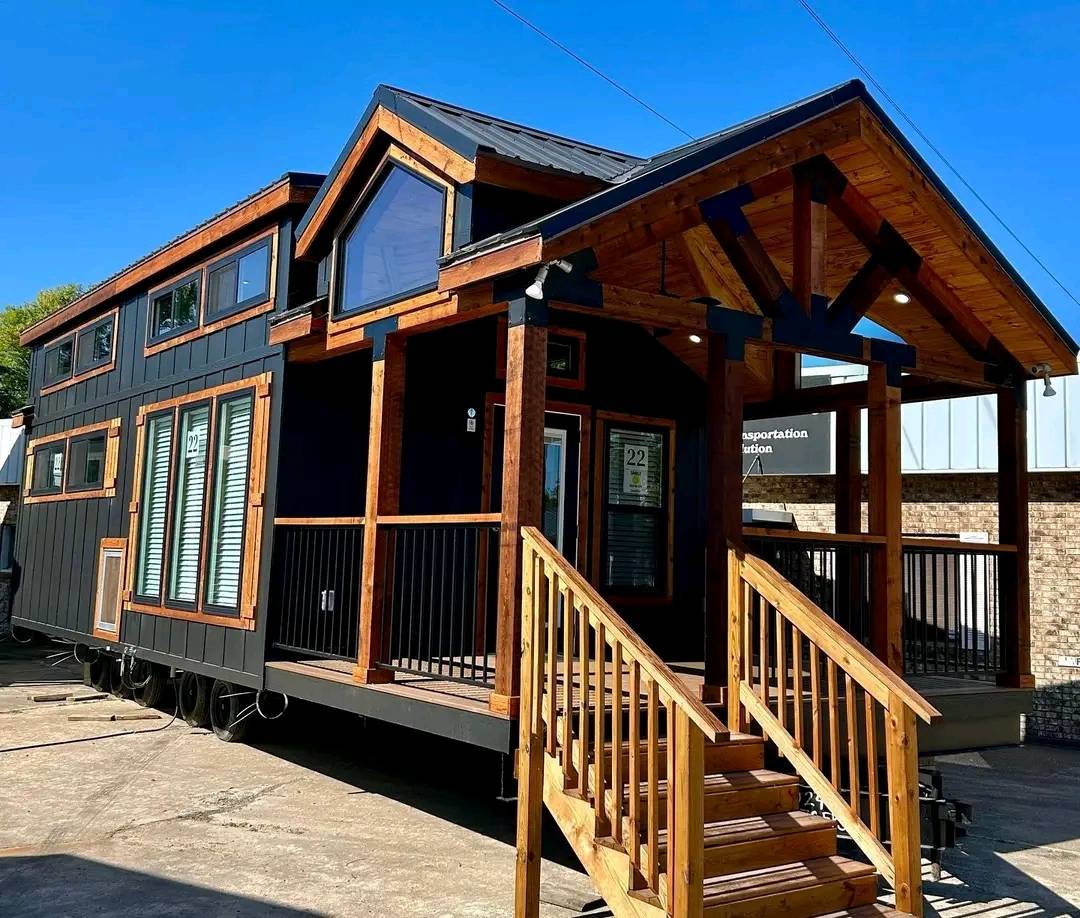A modern tiny house on wheels with dark exterior siding, wooden trim, and large windows. It features a sloped roof and wooden porch under a clear blue sky.