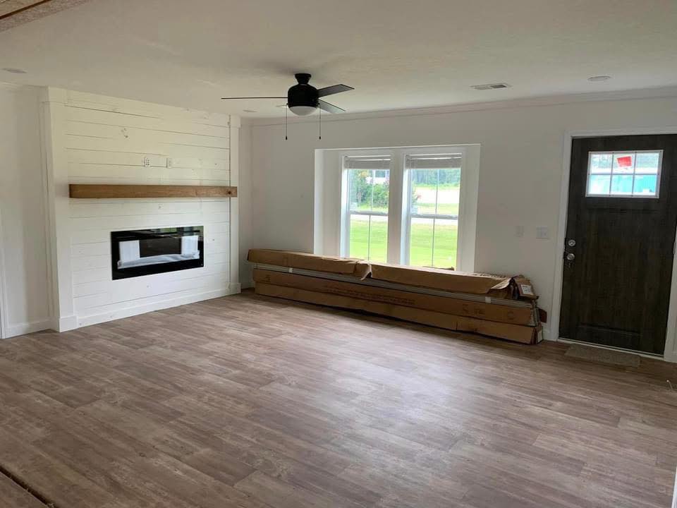 Spacious living room with wood flooring, white shiplap walls, and a modern fireplace. Large windows and a dark wooden door add charm.