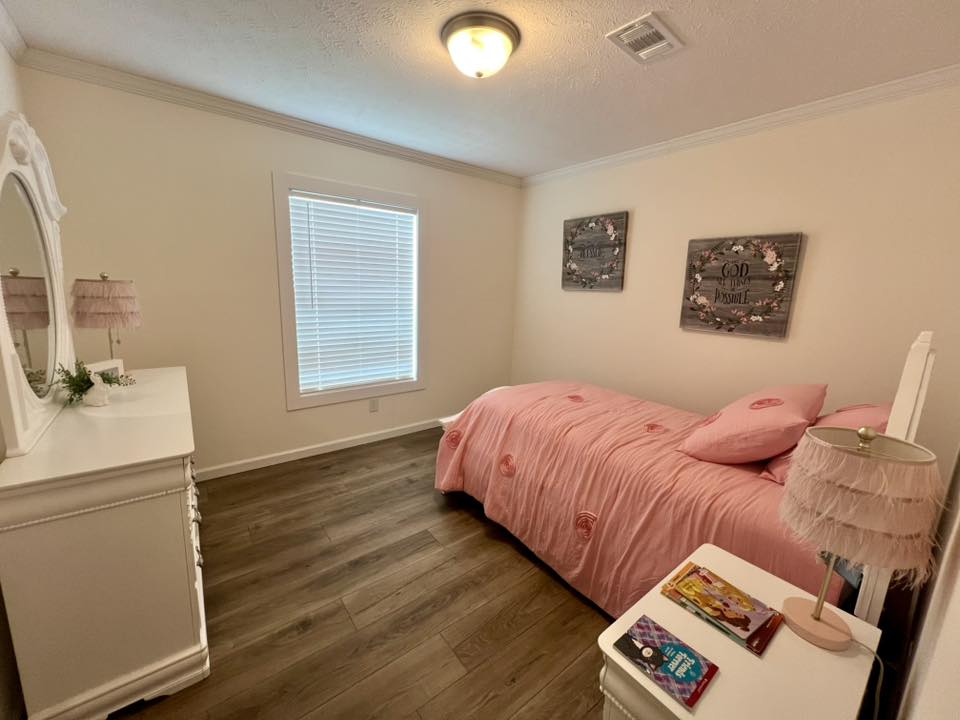A cozy bedroom with a pink-themed decor. A bed with a pink duvet and pillows is against a wall with two floral artworks. A dresser and lamp complete the calm, inviting space.