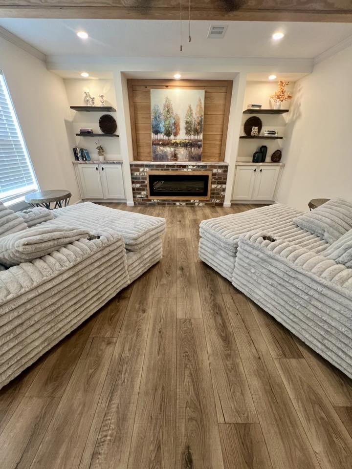 Cozy living room with two plush, light gray recliners facing a faux fireplace. Wooden floors, soft lighting, shelves with decor, and landscape art.