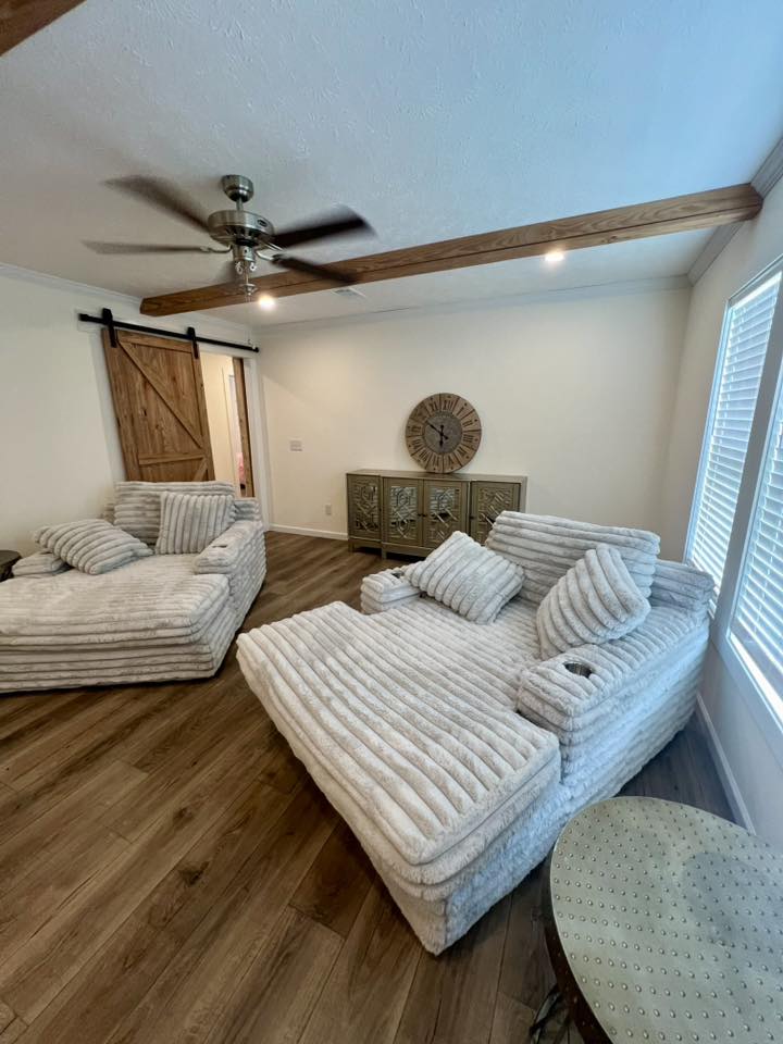 A cozy living room with plush, striped beige seating and matching cushions. A wooden sliding door, a ceiling fan, and a decorative clock create a warm, inviting atmosphere.