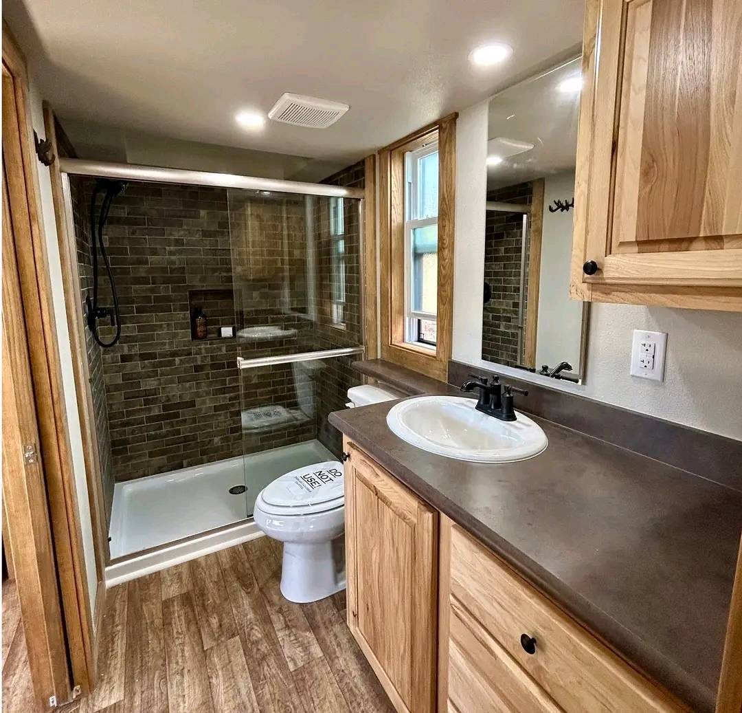 Small modern bathroom with wood accents, featuring a shower with glass doors, brown tiled walls, wooden cabinets, and a white sink. Cozy and warm ambiance.