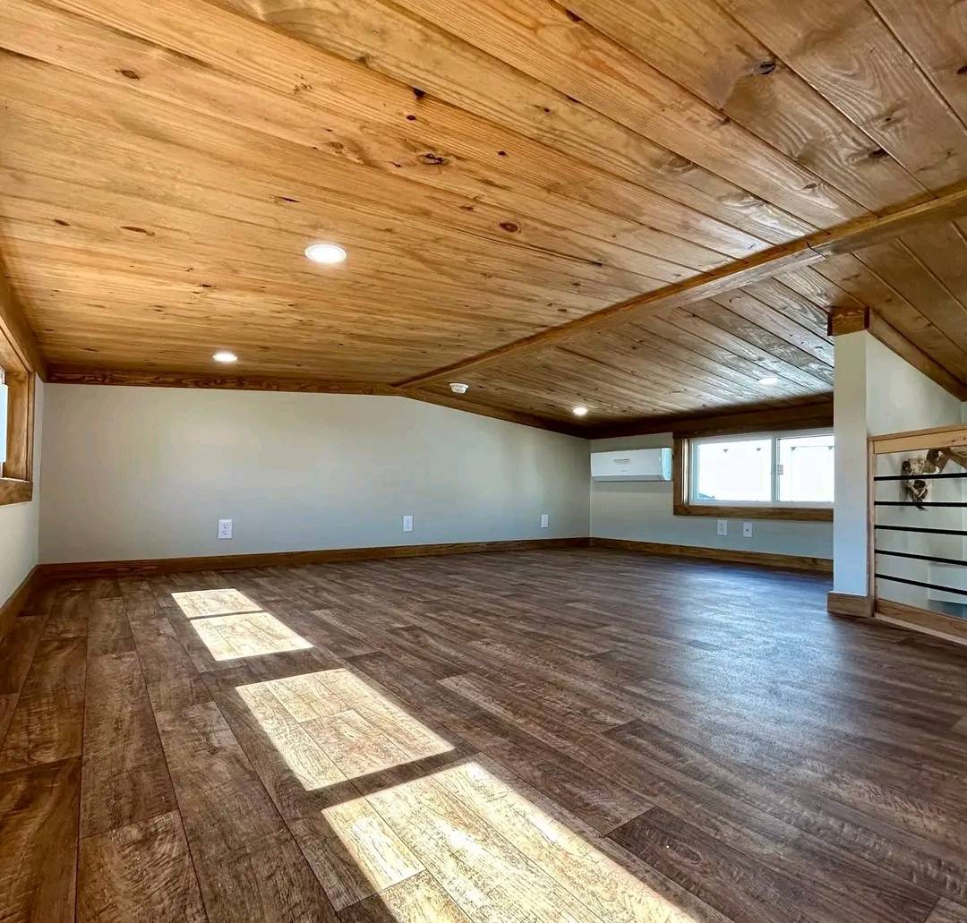 Cozy attic with warm wooden floors and ceilings, illuminated by sunlight from windows. The space feels rustic and inviting, with a simple, uncluttered design.