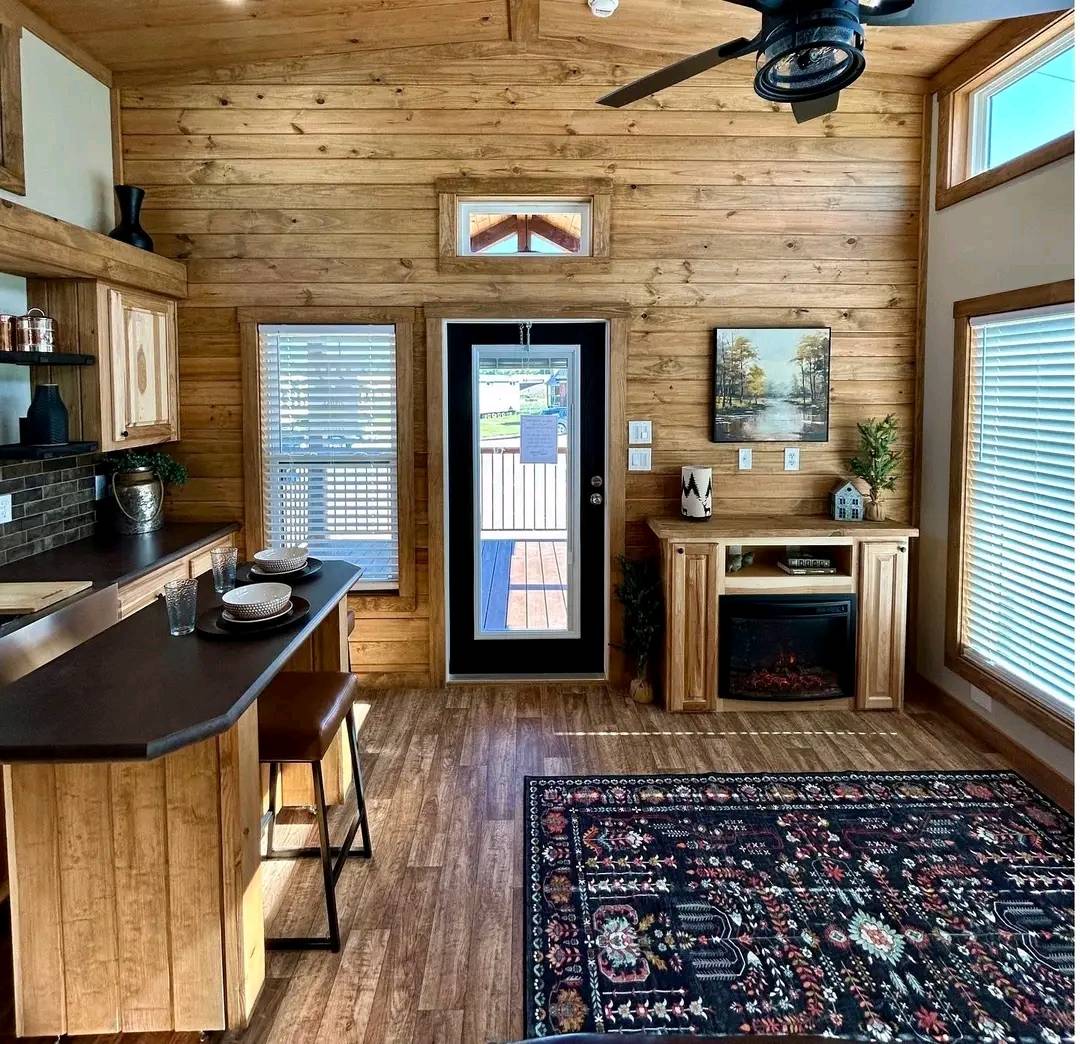 Cozy cabin interior with wood paneling, featuring a small kitchen, a decorative fireplace, and a patterned rug. Sunlight streams through large windows.