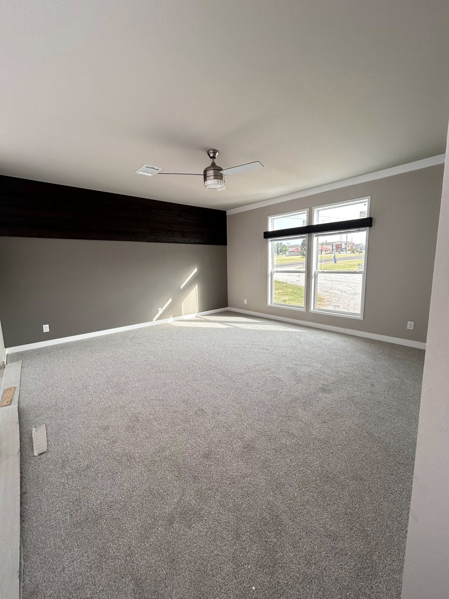 Spacious, empty room with gray carpet and neutral walls. A large window provides natural light, and a modern ceiling fan hangs from the ceiling. Calm and serene ambiance.