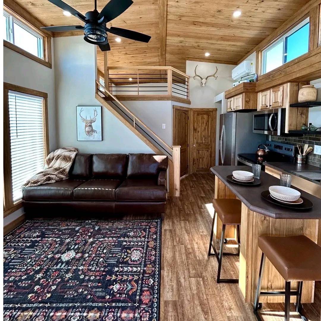 Cozy cabin interior with a leather sofa, patterned rug, and wooden loft. Modern kitchen island with stools, rustic decor, and antler wall art. Warm ambiance.