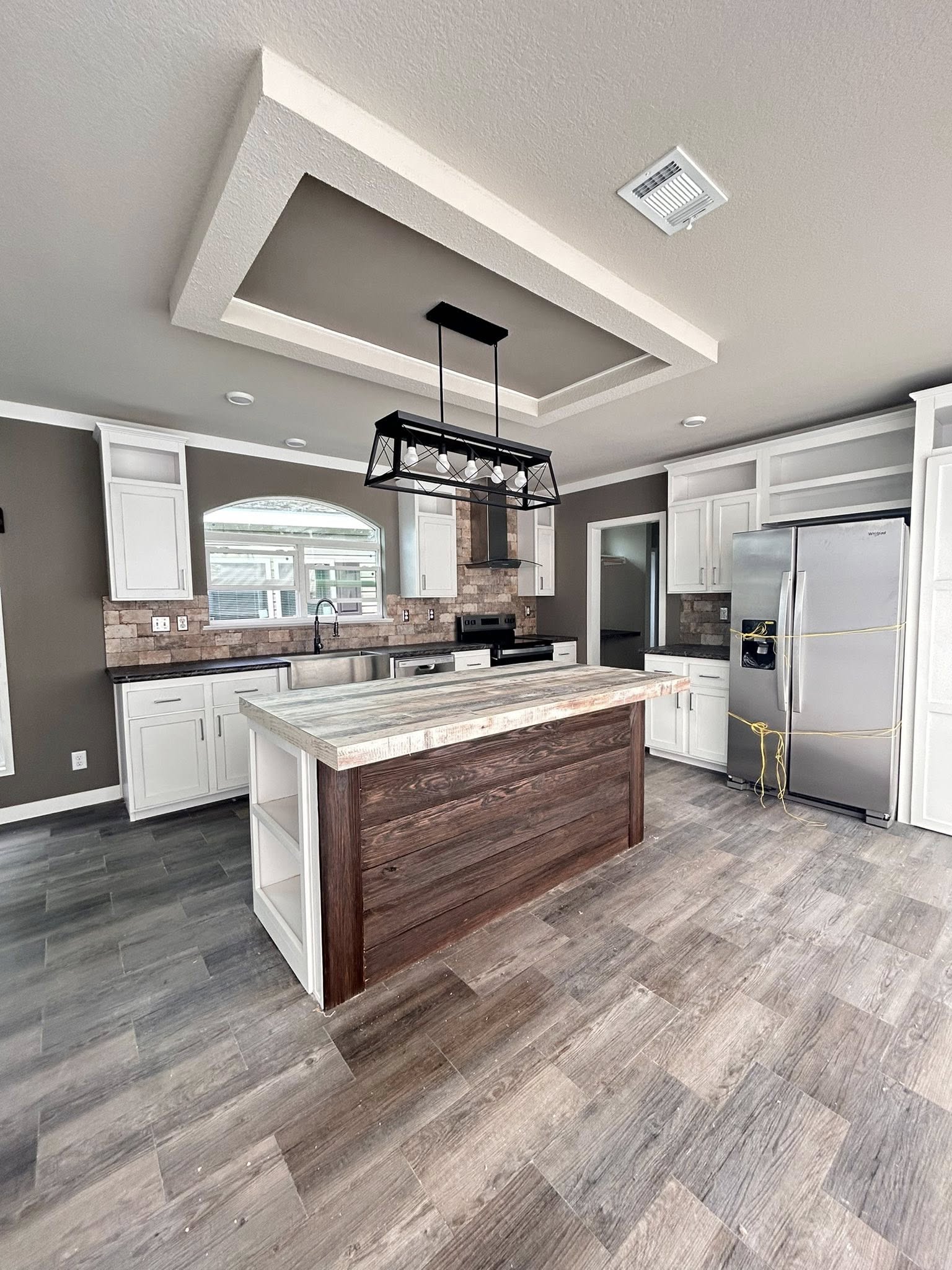 Modern kitchen with a wooden island, distressed wood flooring, stainless steel fridge, white cabinets, and a geometric light fixture. Bright and inviting.