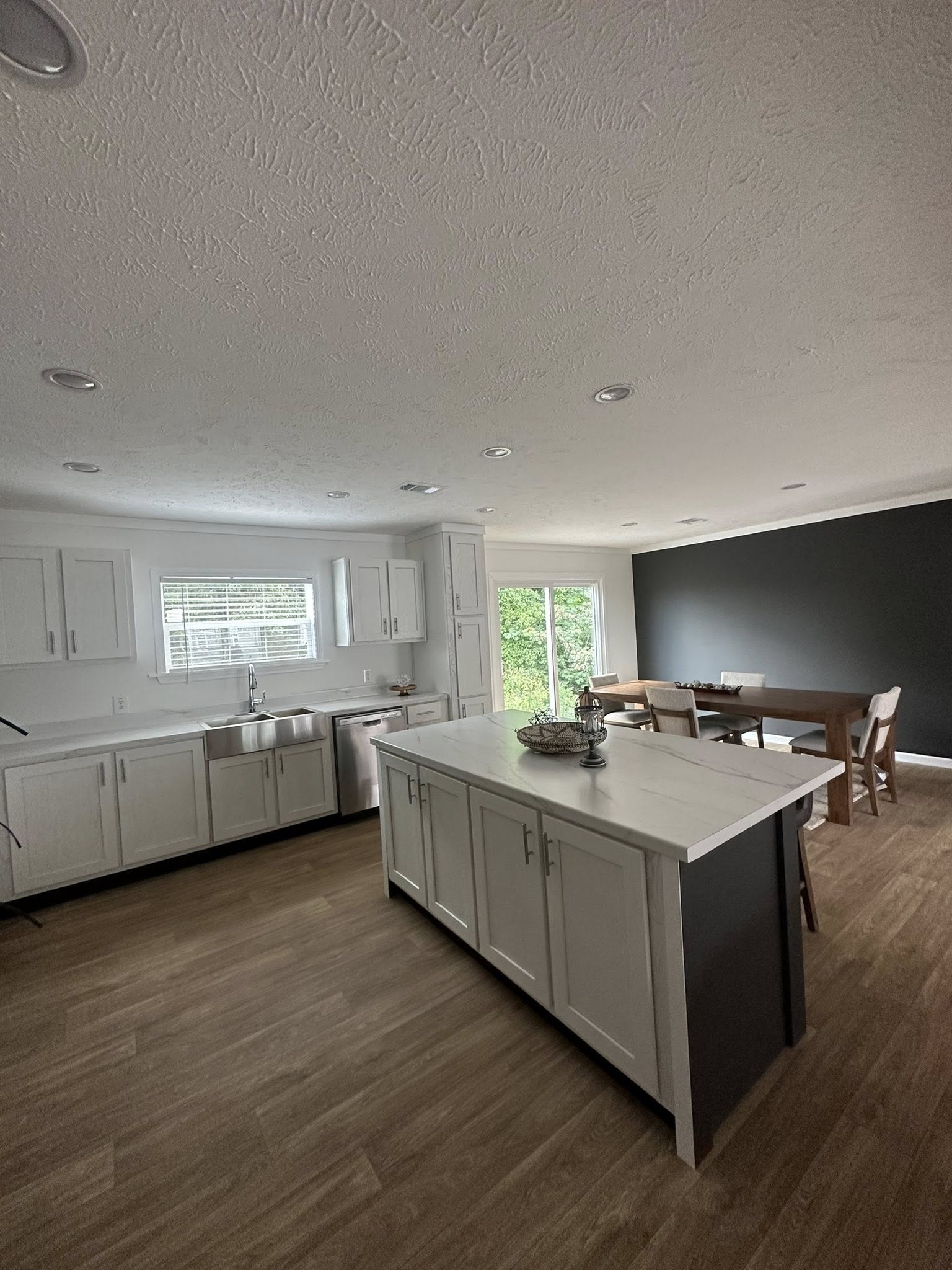 Modern kitchen with white cabinets, stainless steel appliances, and a large island on wood flooring. Adjacent dining area has a dark accent wall.