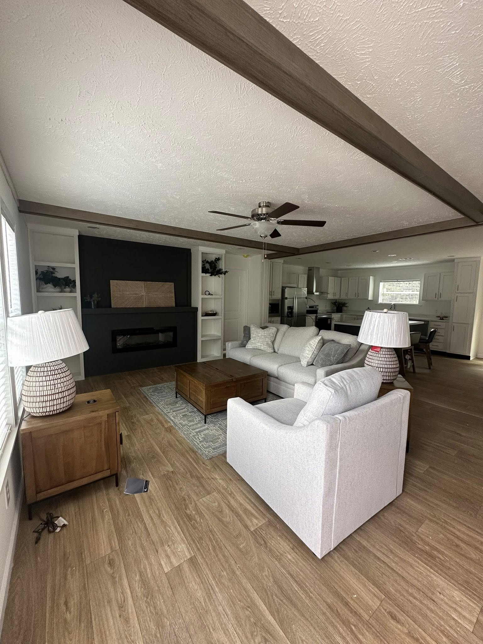Spacious living room with light gray sofas, wooden coffee table, and table lamps. A black fireplace is central on the far wall, under a ceiling fan.