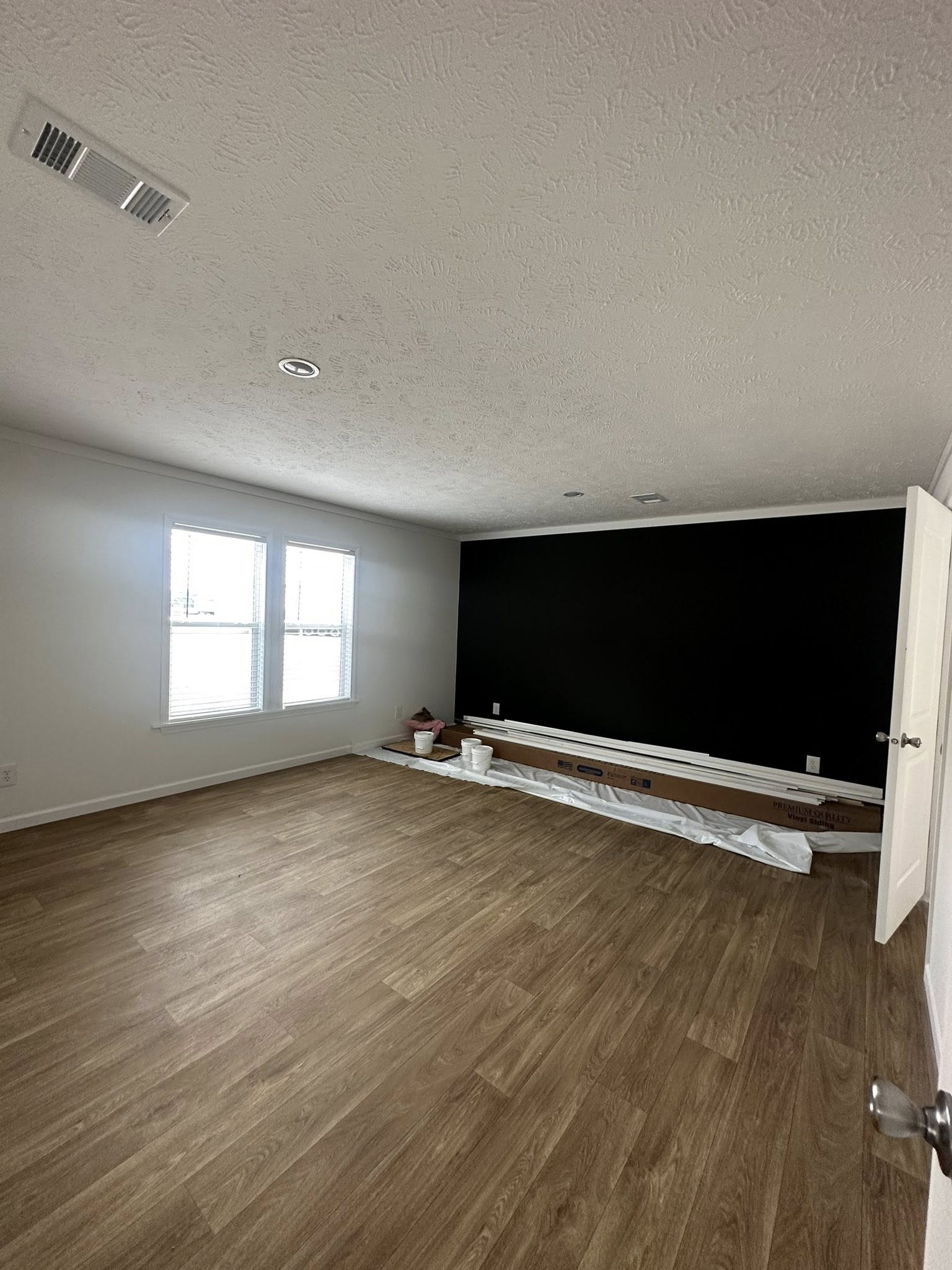 Spacious room with light wood flooring, white walls, and a dark accent wall. Two windows on the left, painting supplies scattered on floor.