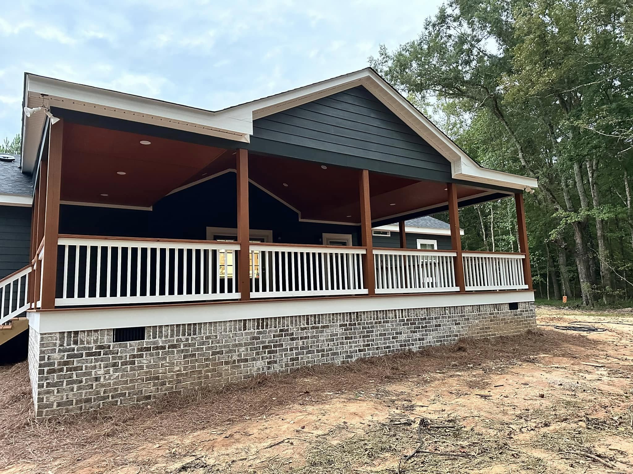Single-story house with dark siding, white trim, and a brick foundation. It has a wooden porch with railings, set amidst trees and a dirt path.