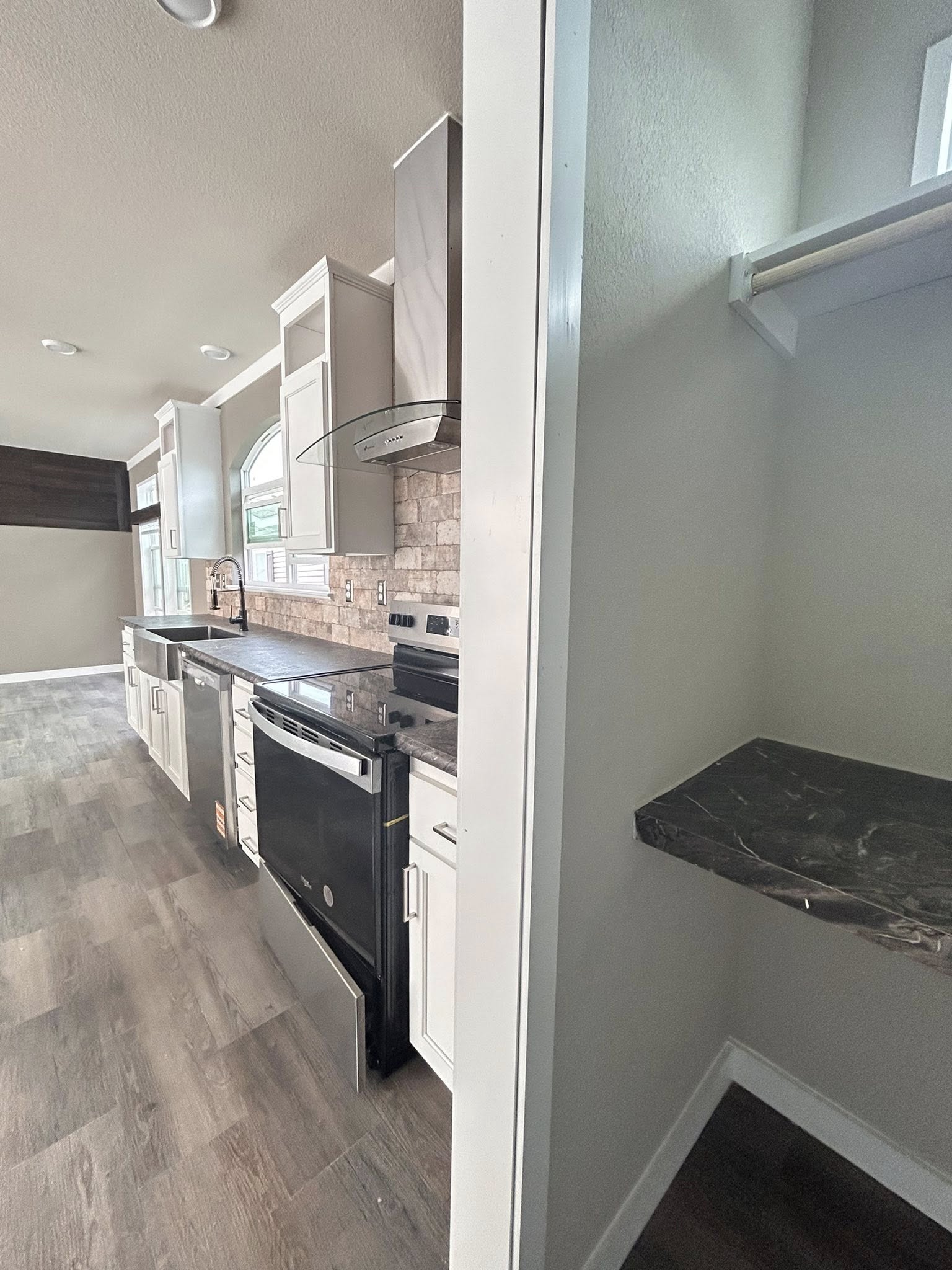 Modern kitchen with white cabinets, stainless steel appliances, and marble countertops. Adjacent to a small pantry with a dark shelf. Bright atmosphere.