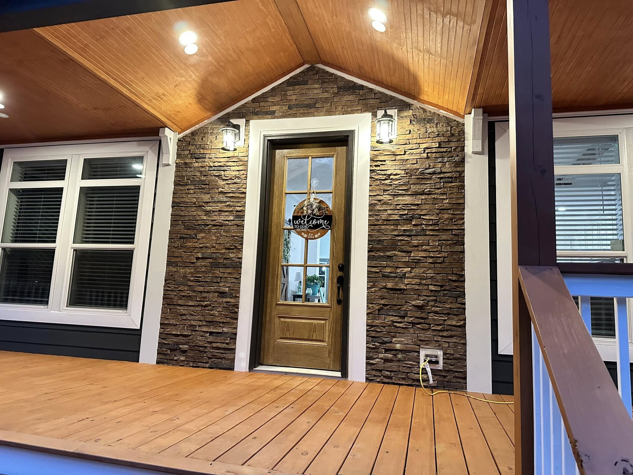 Spacious covered porch with warm wood ceiling and floor, illuminated by recessed lights. It features dark siding and double French doors. Cozy and inviting.