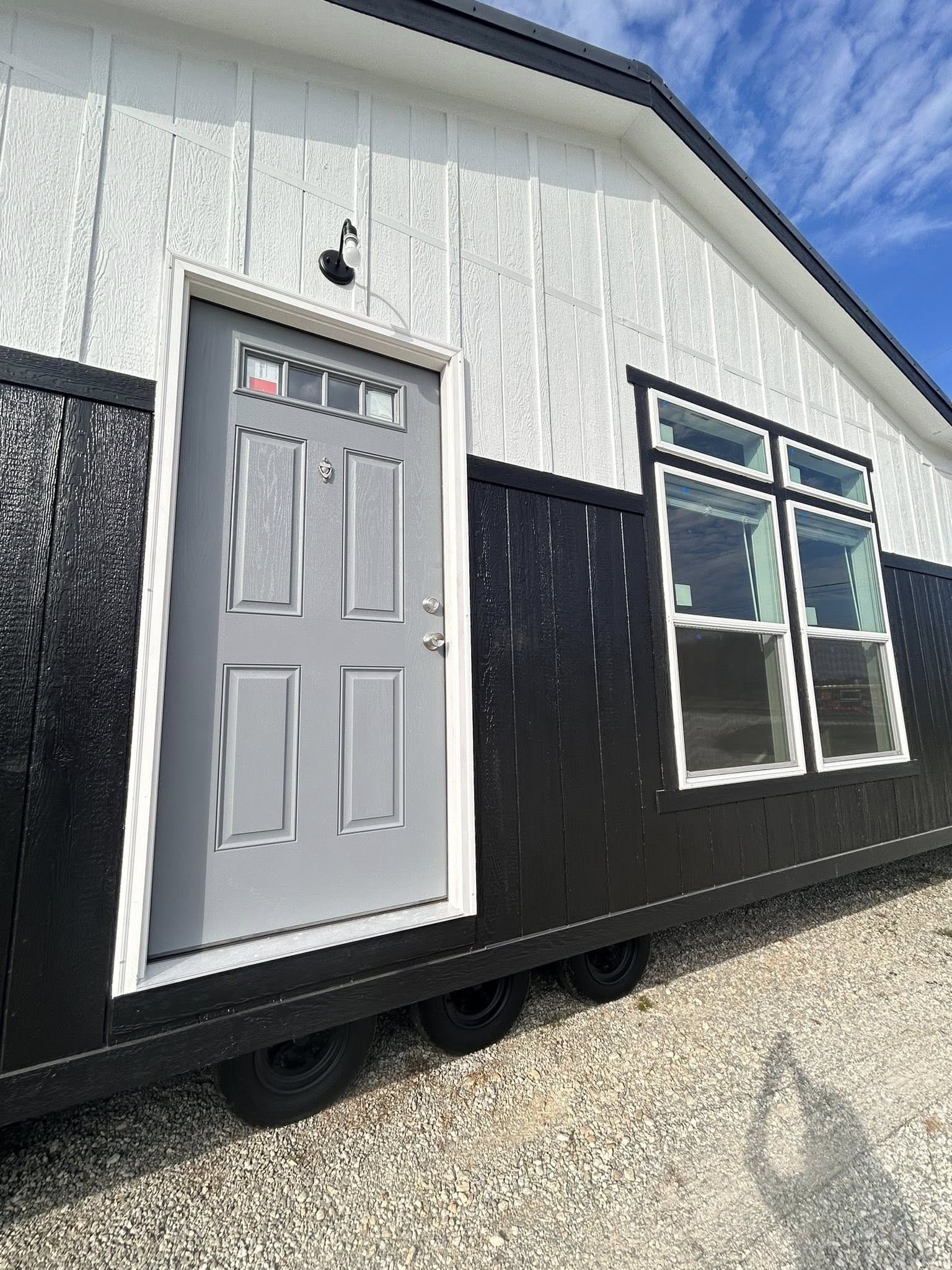 Exterior view of a black and white tiny house with a gray door and large windows, set on wheels on a gravel surface under a blue sky.