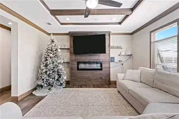 Bright living room with a frosted Christmas tree, white sectional sofa, and a wall-mounted TV above a fireplace. Cozy, festive ambiance.