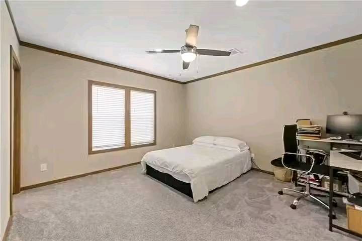 A tidy bedroom with beige walls features a double bed with white linens, a window with blinds, and a ceiling fan. A desk with a chair and computer sits in the corner, creating a calm and functional atmosphere.