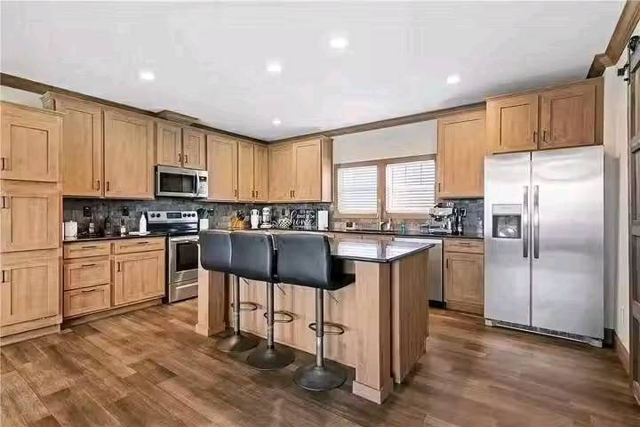 Spacious kitchen with wooden cabinets, stainless steel appliances, and a central island with black leather stools. Bright and modern ambiance.