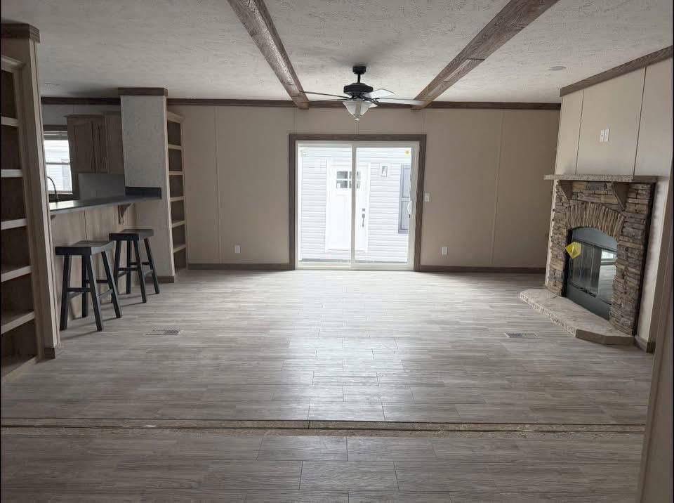 Spacious empty living room with light wood flooring, a stone fireplace on the right, built-in shelves on the left, and a fan above, creating a cozy atmosphere.
