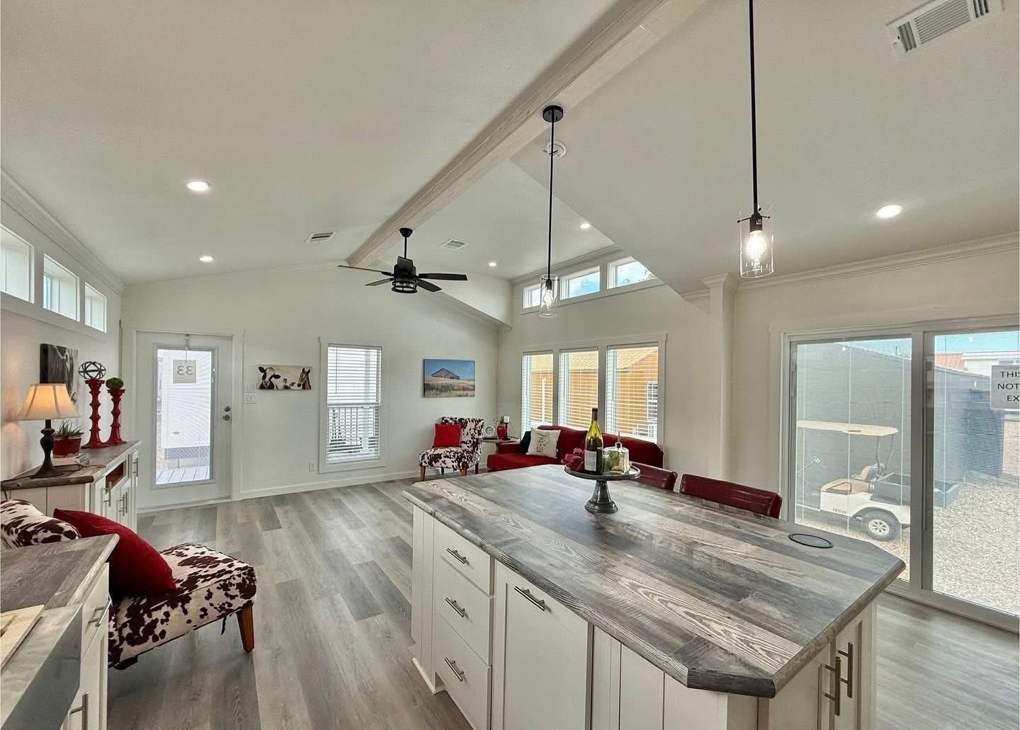 Bright living space with high ceilings, white walls, and light wood flooring. Features modern kitchen island, red furniture, and large windows. Cozy and inviting.