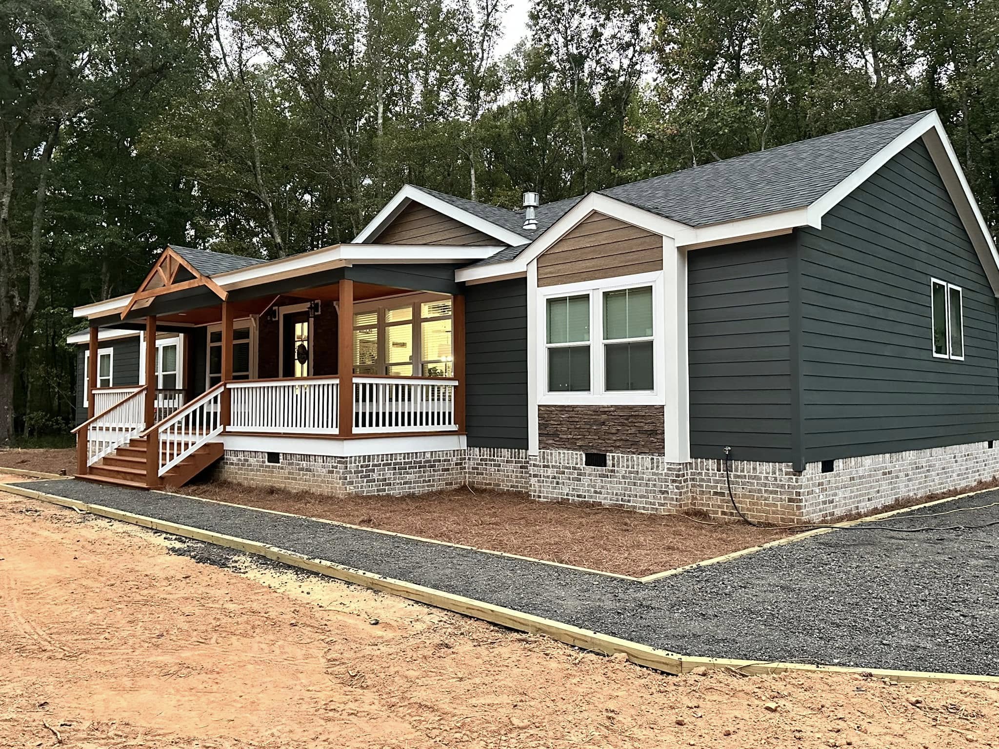Single-story house with a gray exterior, white trim, and wooden porch. The setting is a wooded area with a gravel path, conveying a serene rural atmosphere.