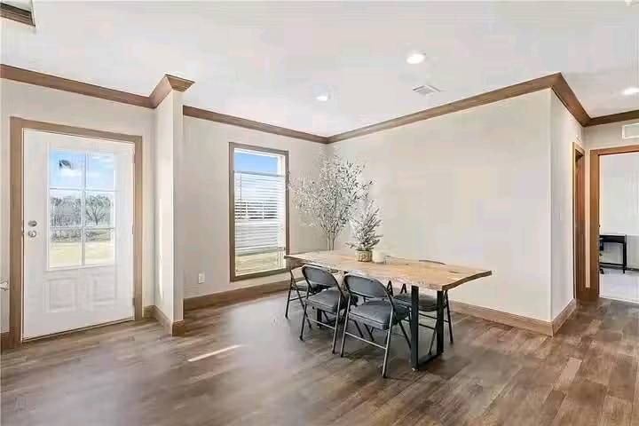 Bright dining area with wooden floors, a simple wooden table, and four dark chairs. A potted plant sits on the table. Natural light streams through a window and glass door, creating a welcoming atmosphere.