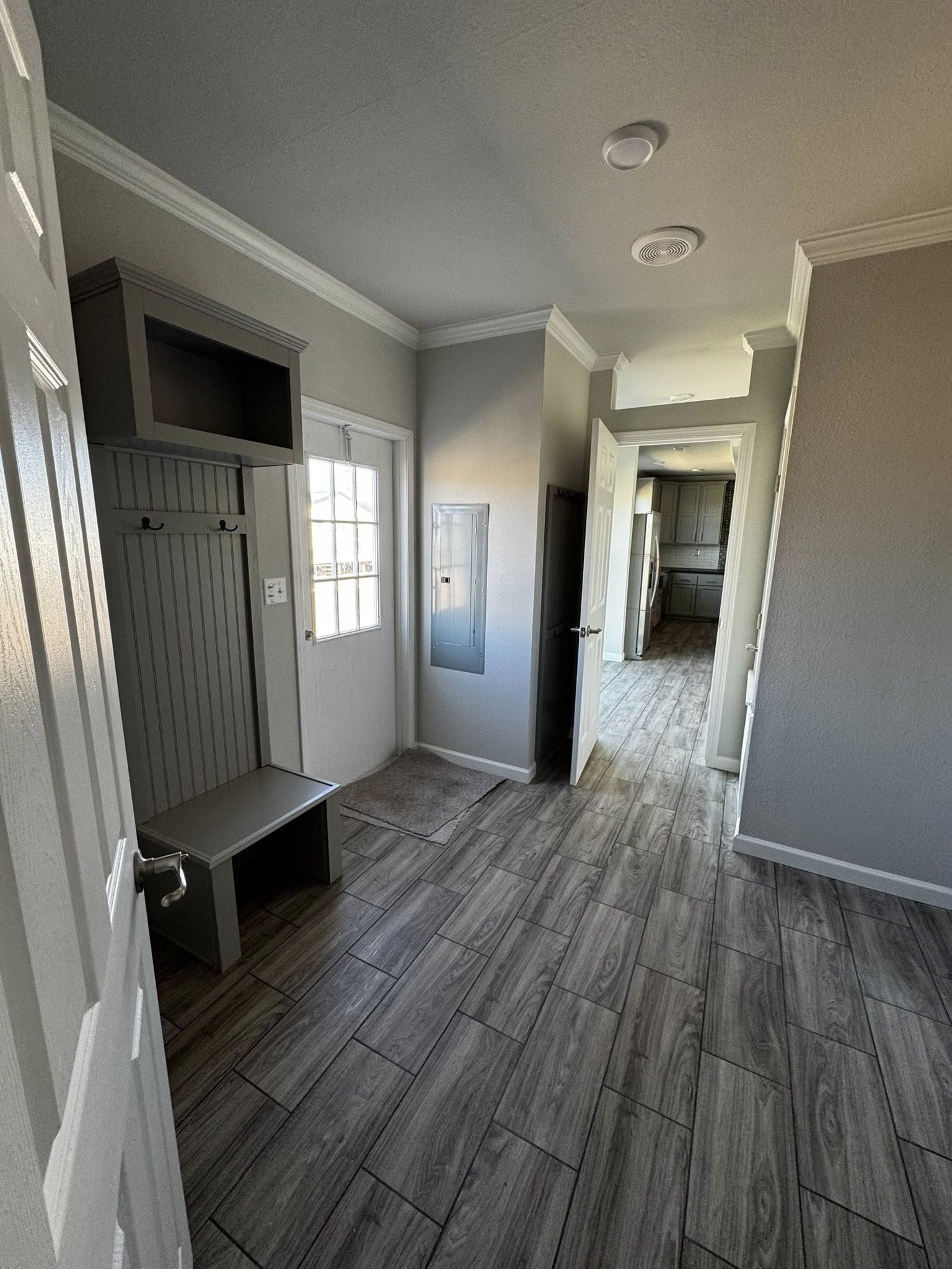 Narrow hallway with gray tile floor leads to a bright kitchen. Left wall has window, bench, hooks. Neutral tones create a calm atmosphere.