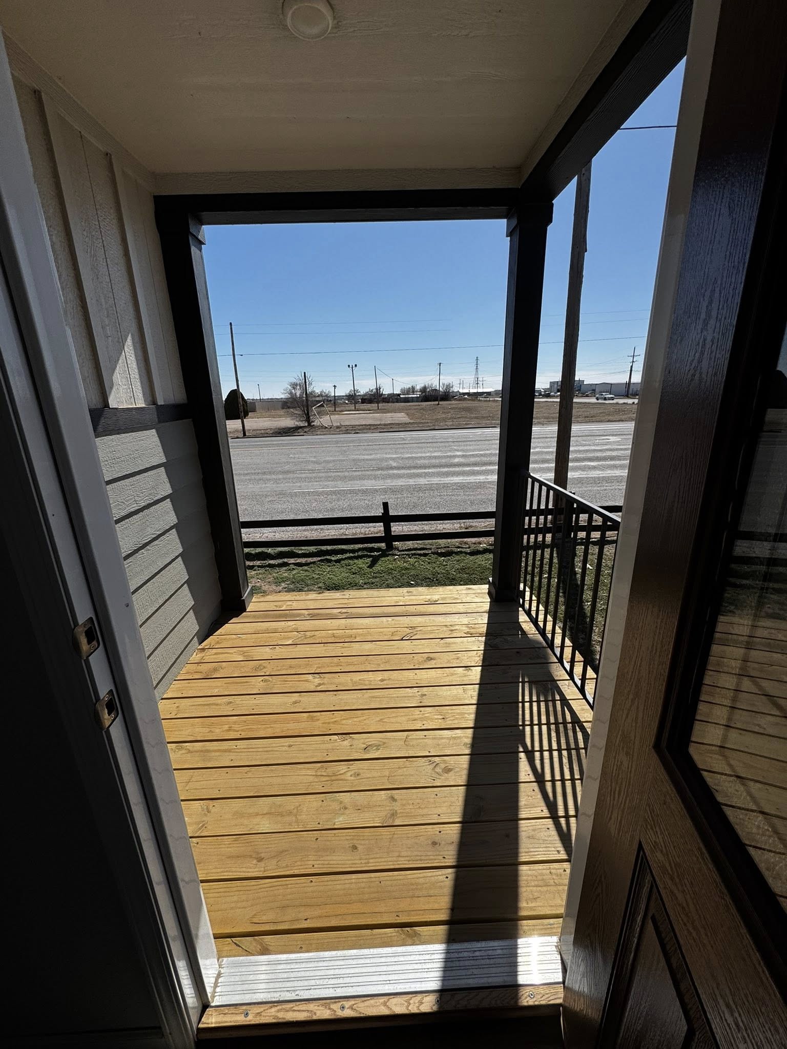 View from an open door onto a small wooden porch with a railing, overlooking a road and an open field under a clear, sunny blue sky.
