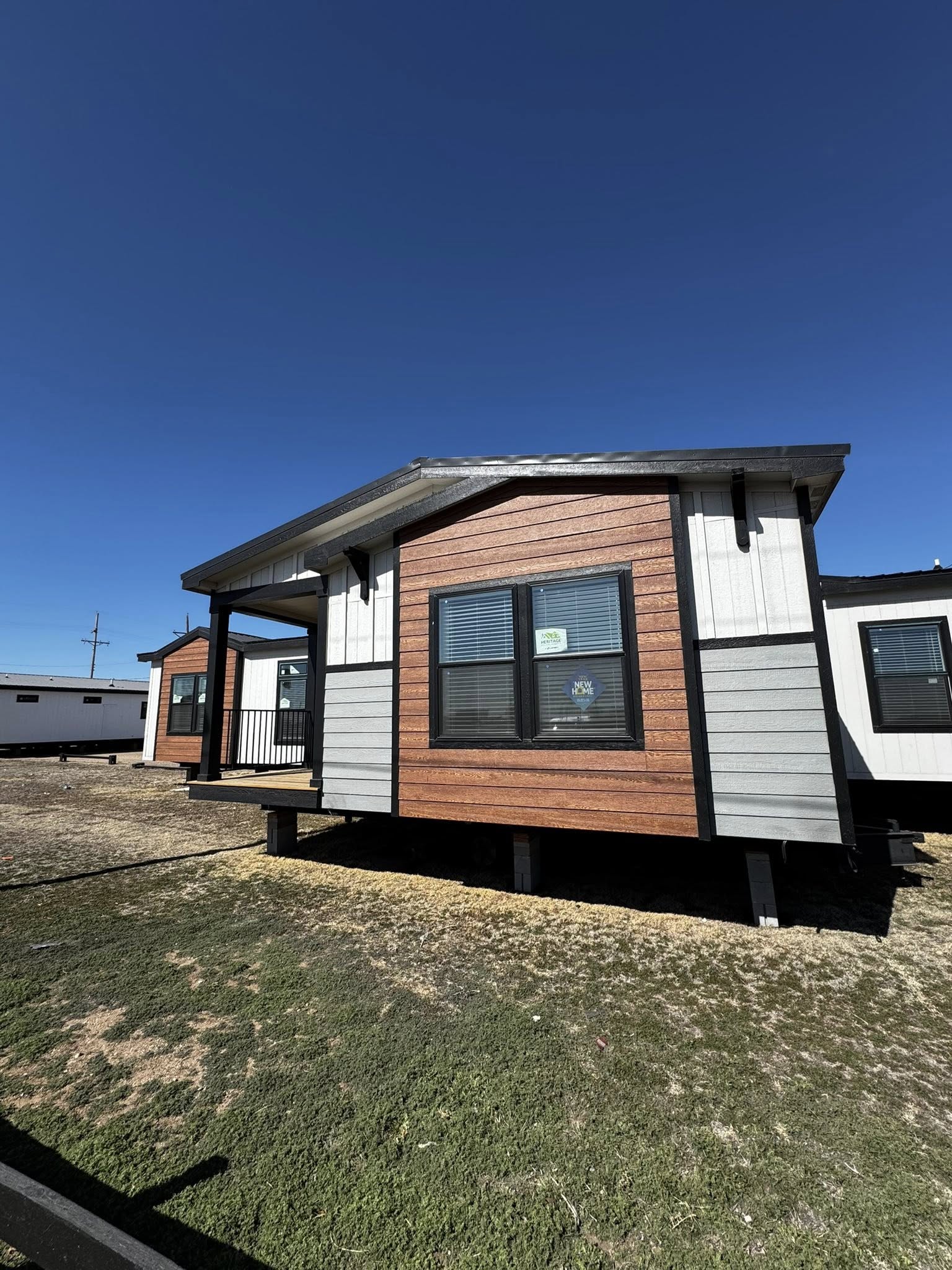 Small modular home on a grassy lot under a clear blue sky. The house features wood paneling and large windows, conveying a modern, cozy feel.