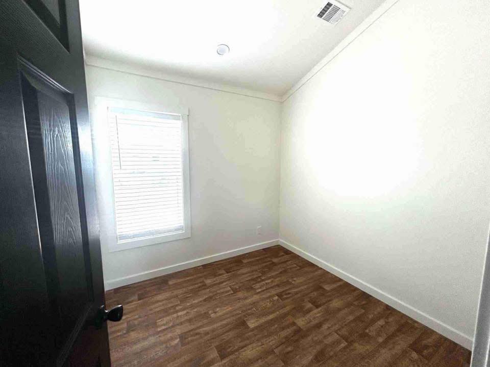 An empty room with white walls and a single window with closed blinds. The floor is wood-patterned, and a black door is slightly open, creating a minimalist and quiet atmosphere.