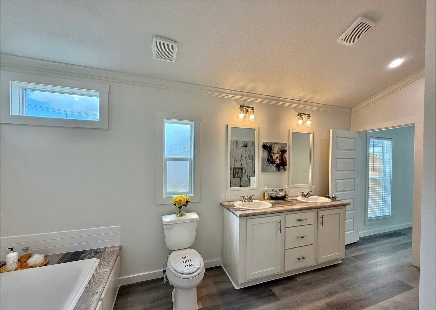 Bright bathroom featuring a double sink vanity with two mirrors, modern lighting, and a decorative cow picture. A "Do Not Use" sign is on the toilet.