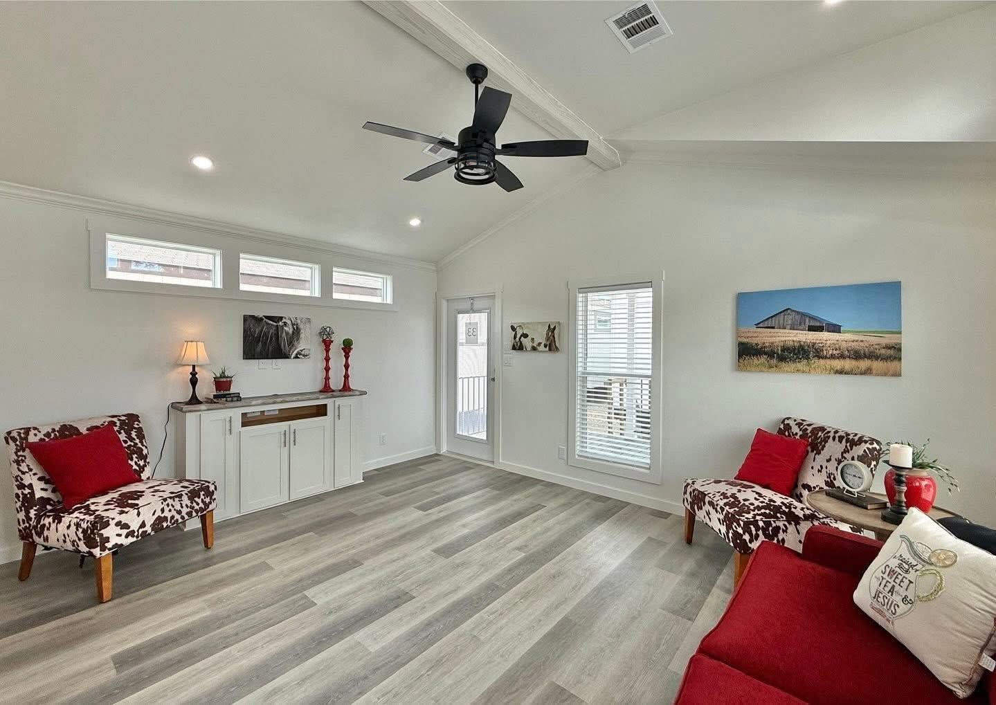 Spacious living room with light wood flooring and white walls. Features floral chairs, red sofa, abstract art, ceiling fan, and warm lighting. Cozy and modern.