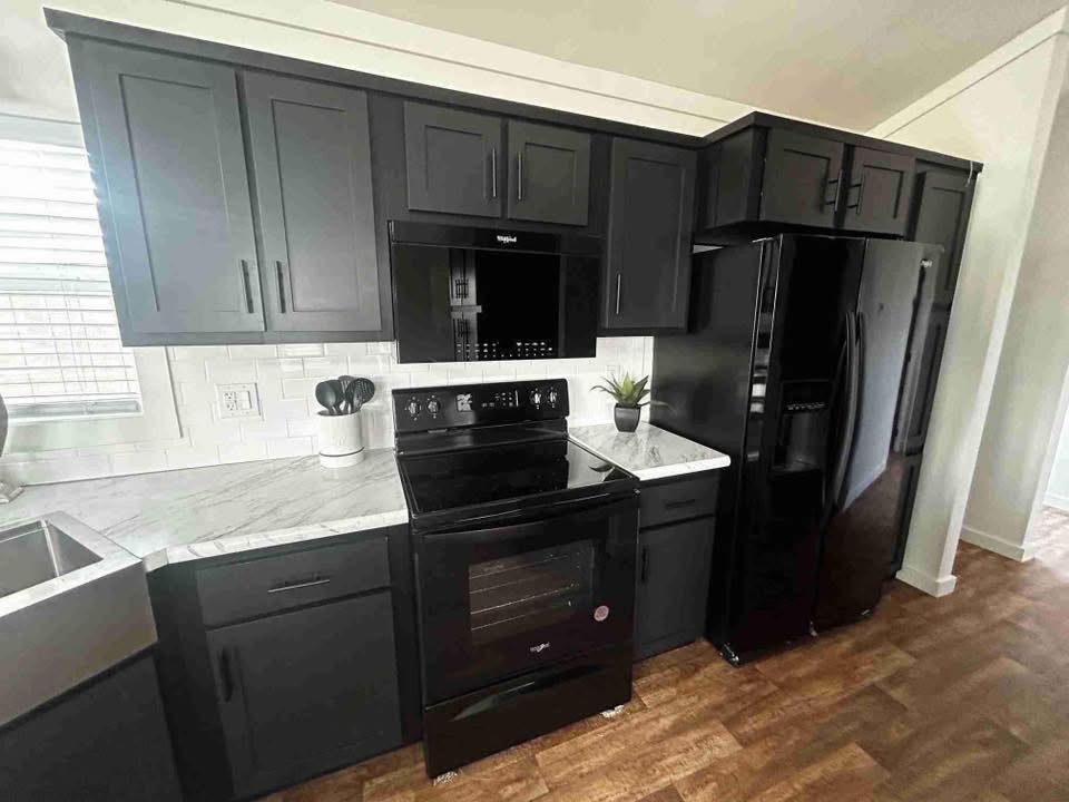 Modern kitchen with sleek black cabinets and appliances, including a stove, microwave, and fridge. Light marble countertops and wood floor create contrast.