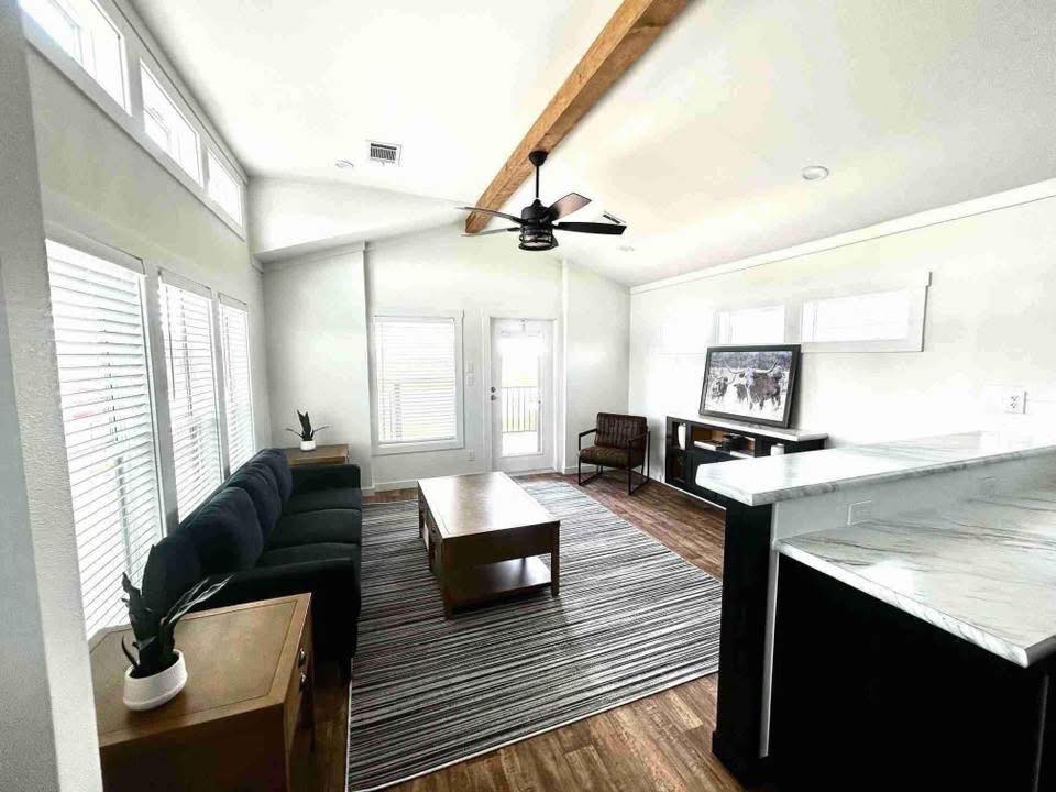 Modern living room with high ceiling, wooden beam, and fan. Features a dark sofa, striped rug, coffee table, and a large window, creating an airy feel.