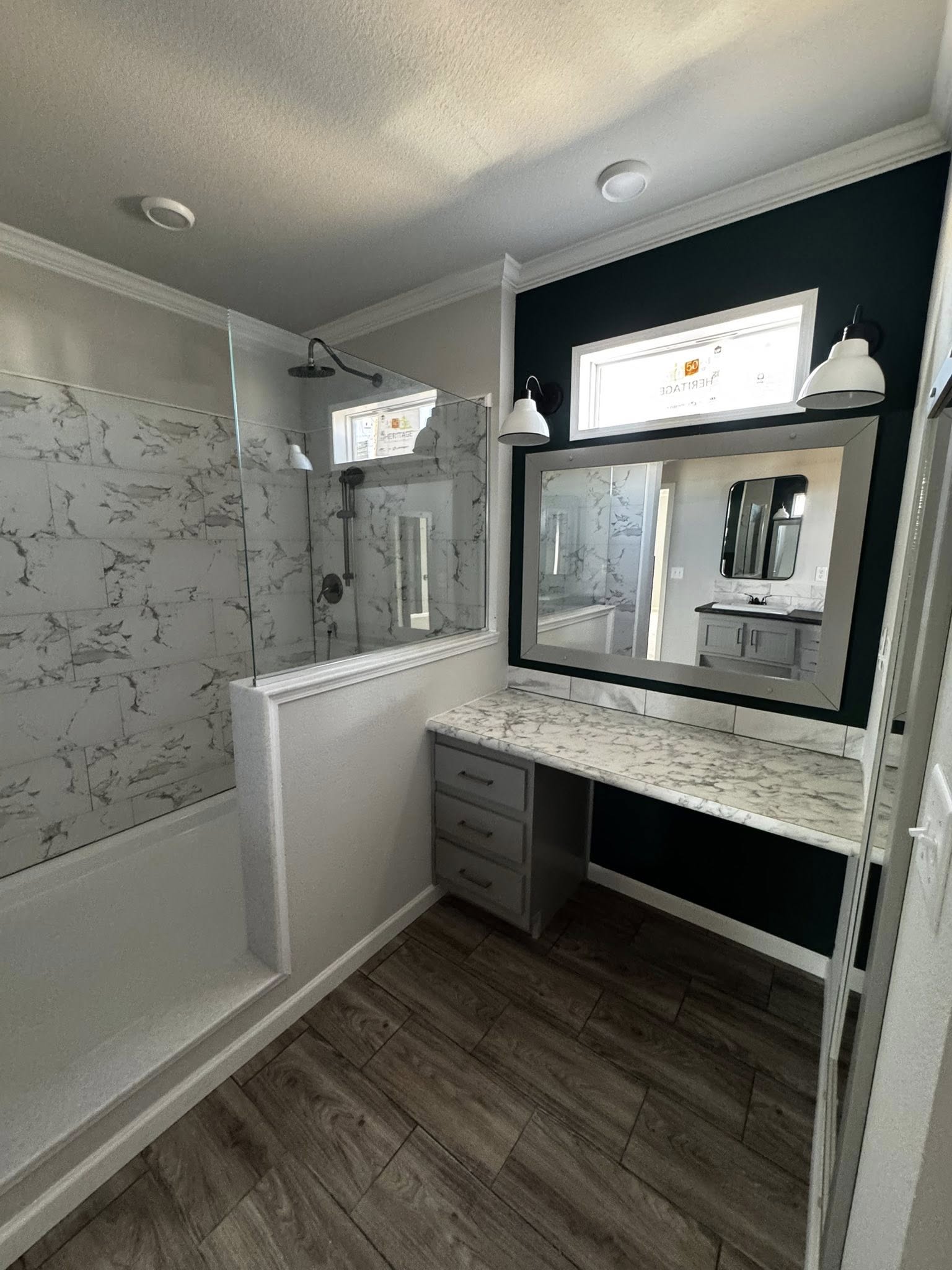 Modern bathroom with marble-tiled shower, gray vanity with large mirror, dual light fixtures, and wood-patterned floor. Bright and elegant atmosphere.