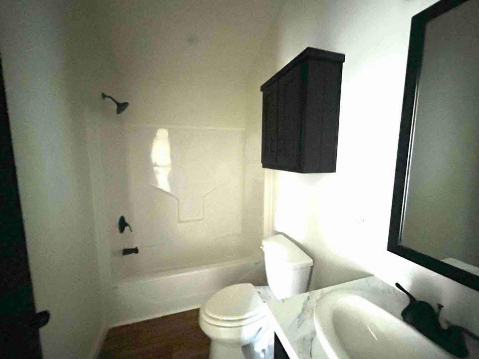 A small bathroom with a bathtub-shower combo, dark wood cabinet above the toilet, white sink with marble-like countertop, and a large mirror.