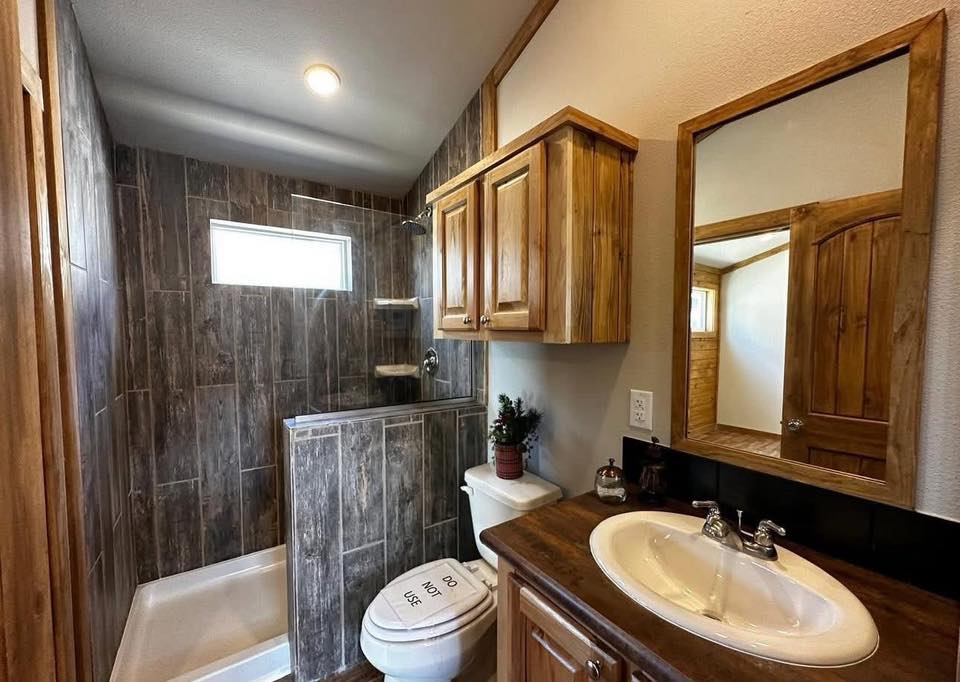 A cozy bathroom features a wooden vanity with a white sink, a large mirror, and a rustic cabinet. The shower has dark brown tiles and a small window, creating a warm, inviting ambiance.