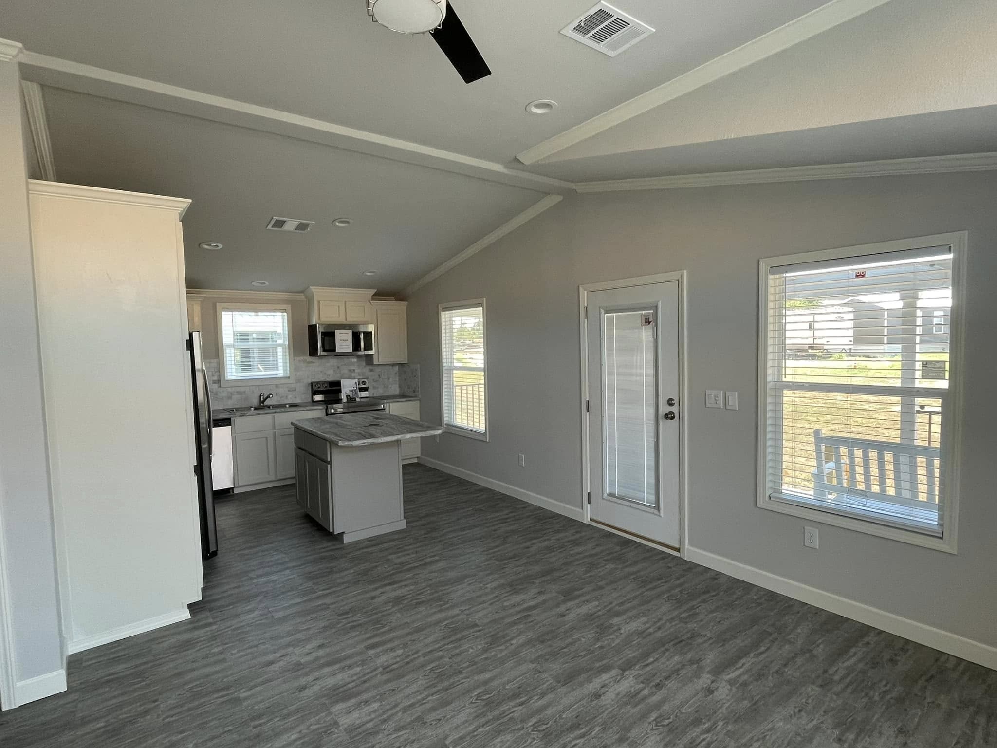 Modern kitchen and living area with gray flooring and light walls. Features include white cabinets, stainless steel appliances, and large windows.
