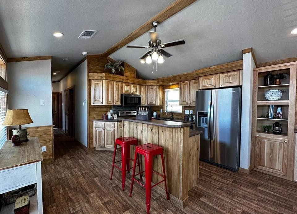 Spacious kitchen with wood cabinetry and floors, featuring stainless steel appliances and a central island with red stools. Ceiling fan overhead. Cozy ambiance.