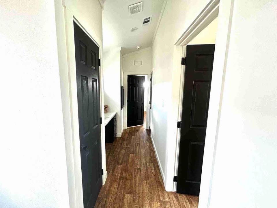 A narrow hallway with light walls and wood flooring features multiple black doors on both sides. Soft natural light streams in, creating a calm atmosphere.