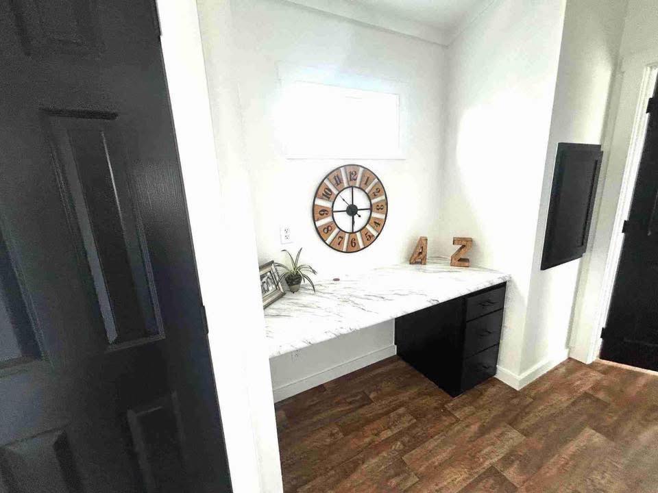 A small alcove with a white marble desk, black drawers, and wall clock. Decor includes small plants and wooden letters. The space is minimal and modern.