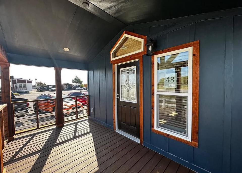 Porch view with dark wood paneling, a glass-paneled door, and window marked "43." Sunlight casts shadows, while parked cars and buildings are visible outside.