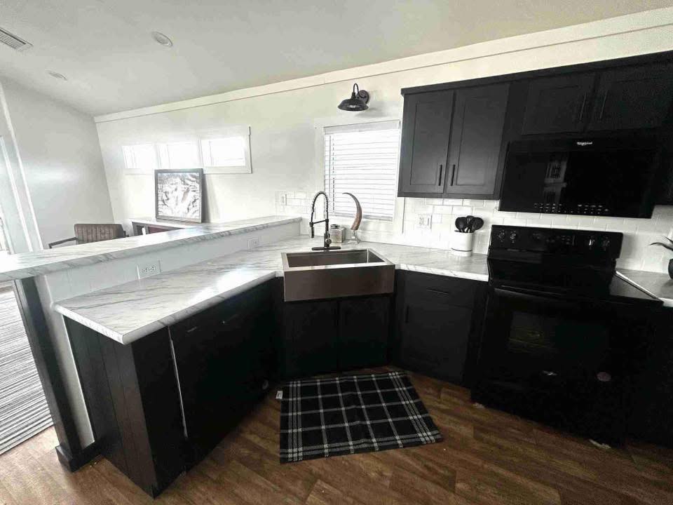 Modern kitchen with dark cabinetry and marble countertops. Features a stainless steel farmhouse sink, black appliances, and wooden flooring. Bright, airy ambiance.