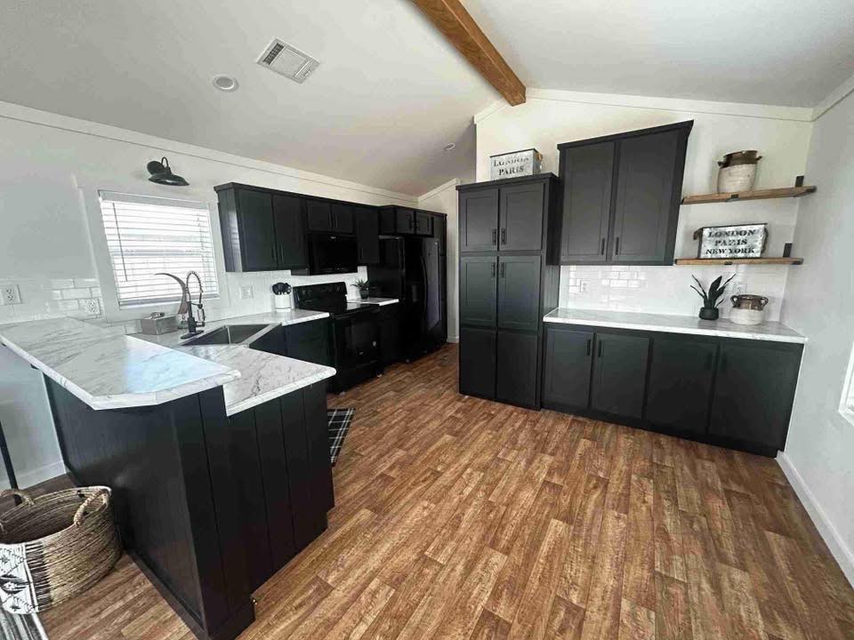 Modern kitchen with black cabinets, white countertops, and wood flooring. Features open shelving, a marble-topped island, and stainless steel faucet. Cozy and sleek design.
